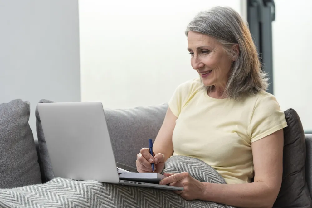 Ältere Frau sitzt mit Laptop und Notizen auf dem Sofa